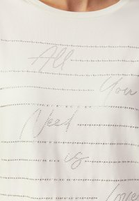 Weißes T-Shirt mit kurzen Ärmeln, das mit silbernem Strasstext und horizontalen Linien verziert ist. Weicher Stoff mit glatter Textur und lässiger Passform.