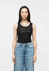 Jeune femme portant un haut noir sans manches avec le logo "Guess" en strass et un jean bleu taille haute à jambes larges, debout devant un fond uni.