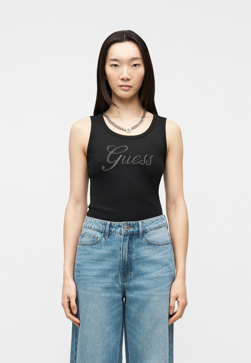Jeune femme portant un haut noir sans manches avec le logo "Guess" en strass et un jean bleu taille haute à jambes larges, debout devant un fond uni.