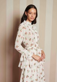 Φλοράλ φόρεμα σε ελαφρύ, διάφανο ύφασμα. Διαθέτει ριγέ κολάρο και μανίκια, μέση πεπλούν και ζώνη που δένει. Ανοιχτό ροζ φόντο.