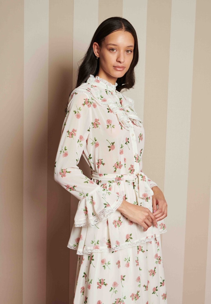 Φλοράλ φόρεμα σε ελαφρύ, διάφανο ύφασμα. Διαθέτει ριγέ κολάρο και μανίκια, μέση πεπλούν και ζώνη που δένει. Ανοιχτό ροζ φόντο.