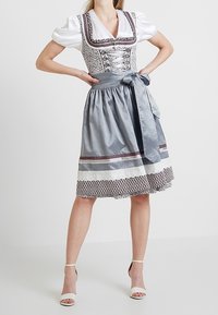 Kobieta ubrana w tradycyjną szaro-białą sukienkę dirndl z bufiastymi rękawami i zawiązanym fartuszkiem, stoi z rękami na biodrach.