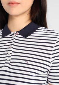 Kobieta ubrana w biało-granatową koszulkę polo w paski z granatowym kołnierzykiem i białymi guzikami, na której na piersi znajduje się małe haftowane logo.