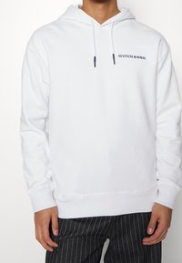 Muž oblečený v bílé mikině s kapucí s nápisem "SCOTCH & SODA" na hrudi a černých kalhotách s proužky, stojící před jednobarevným pozadím.