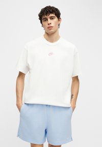 Νέος άνδρας με σγουρά μαλλιά στέκεται με τα χέρια στις τσέπες, φορώντας λευκό t-shirt Nike και ανοιχτό μπλε σορτς Nike μπροστά σε ένα απλό φόντο.