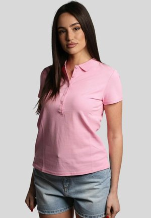 Polo shirt in cotone rosa con colletto, maniche corte e patta dei bottoni. Presenta un logo ricamato discreto e una texture liscia. Indossato con pantaloni corti di jeans.