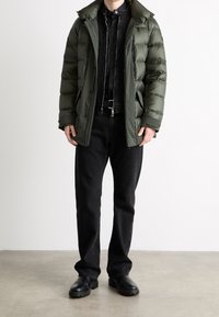 Chaqueta tipo puffer en verde oliva oscuro, diseño acolchado, con cremallera frontal y un cuello alto; combinada con una camisa negra y jeans negros, con botas negras gruesas.