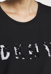 Svart kortärmad skjorta med bokstäverna "DKNY" i växelvis svarta och vita paljetter, med en slät yta och rund halsringning.