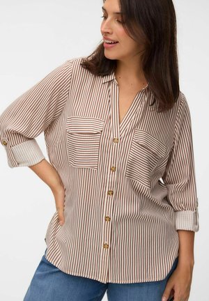 Kvinde iført en brun og hvid vertikalt striped skjorte med opkørte ærmer og blå jeans, ser til siden med et let smil.