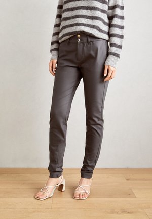 Person iført tætsiddende mørkegrå bukser, stribet grå og sort sweater og hvide sandaler med hæle og stropper, der står på gulv af træ.