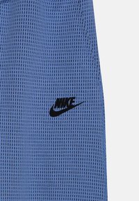 Blå nätshorts med ett rutmönster, med en svart Nike-logotyp på den nedre sidan. Tyget verkar texturerat och lätt.