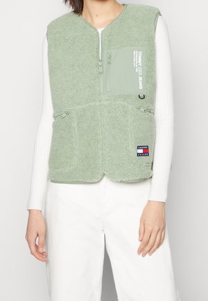 Person iført lysegrøn fleecevest med lynlås og logopatch over en hvid bluse med lange ærmer og hvide bukser, stående mod en ensfarvet baggrund.