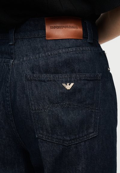 Σκούρα μπλε τζιν από denim διαθέτουν δερμάτινο ταμπελάκι στην πλάτη με ανάγλυφο το "EMPORIO ARMANI". Μικρό λογότυπο σε χρυσό χρώμα στο πίσω τσεπάκι.