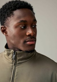 Jachetă verde măsliniu cu fermoar și guler înalt, cu o textură netedă și detalii negre la fermoar, purtată de un bărbat cu părul negru tăiat scurt.