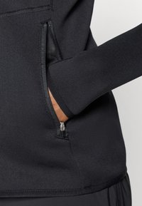 Top sportivo nero con finitura testurizzata, dotato di una tasca con zip sul lato e maniche aderenti. Tessuto liscio con elasticità.