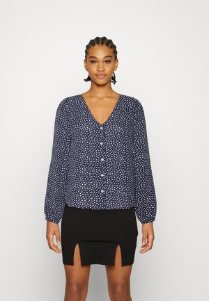 Blusa azul marinho com bolinhas brancas, decote em V, mangas longas em balão e fecho de botão. Combinada com uma saia preta com aberturas laterais.