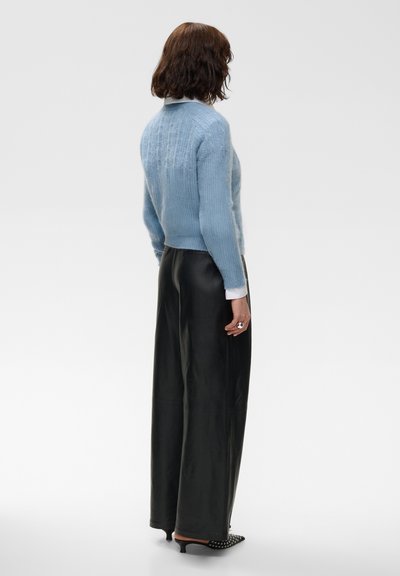 Femme aux cheveux mi-longs portant un pull bleu clair, des poignets de chemise blancs, un pantalon large noir et des talons noirs cloutés, debout de profil.