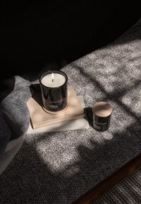 Svart ljusbehållare med vit vax, lågan tänd, på en ljusfärgad fyrkantig underlägg. Liten matchande burk bredvid den på en texturerad filt.