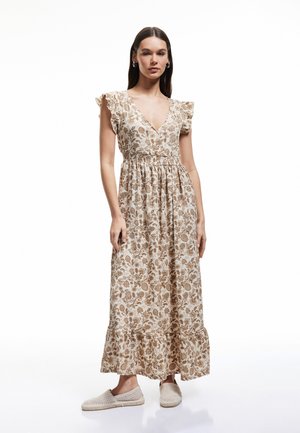 Maxi kjole i creme med brunt blomstermønster, der har en dyb V-udskæring, bølgede ærmer og en samlet talje. Matchet med beige vævede slip-ons.