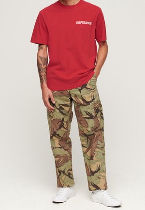 Mann trägt rotes "SUPERDRY" T-Shirt, Camouflage-Cargohose, weiße Sneakers, mit tätowiertem linken Arm, steht mit rechter Hand in der Tasche.