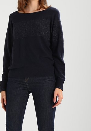 Žena v tmavě modrém svetru s dlouhým rukávem se stříbrnými proužky a tmavě modrých džínách stojí před jednoduchým světlým pozadím.