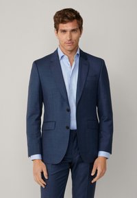 Marineblaues, tailliertes Sakko mit Schlitzen, zwei vorderen Taschen und dunklen Knöpfen, kombiniert mit einem hellblauen Hemd darunter.