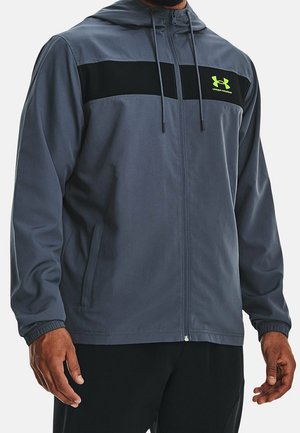 Hombre con chaqueta gris con cremallera y capucha, con una franja negra en el pecho y un logo de Under Armour en neón verde, combinado con pantalones negros.