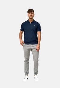 Hombre de pie con la mano en el bolsillo, vistiendo polo azul marino, pantalones deportivos gris claro y zapatillas blancas sobre fondo liso.