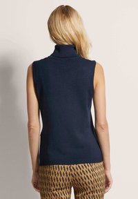 Marineblauer ärmelloser Rollkragenpullover mit geripptem Kragen, glatter Textur und taillierter Silhouette. Kombiniert mit gemusterten Hosen.