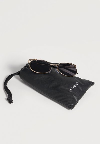 Lunettes de soleil rondes à monture dorée avec des verres foncés reposant sur une pochette noire à cordon portant l’étiquette « Off-White™ » sur une surface blanche.