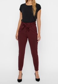 Bordeauxrode taps toelopende broek met een strikband, voorzien van zijzakken. Gecombineerd met een zwart T-shirt met korte mouwen en zwarte hoge hakken.