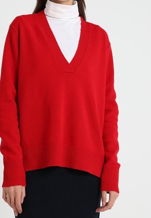 Kvinde iført en knaldrød V-hals sweater over en hvid rullekrave, kombineret med et sort skørt, stående foran en ensfarvet hvid baggrund.