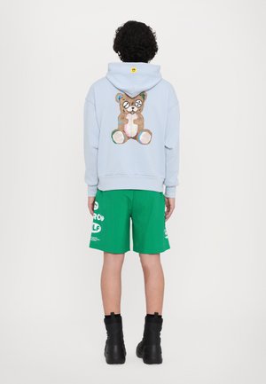 Sudadera azul claro con un diseño de oso de peluche en la parte posterior, combinada con pantalones cortos verdes y botas negras hasta el tobillo.