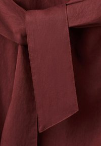 Gros plan d'une ceinture en tissu nouée de couleur rouge foncé sur un vêtement assorti, montrant une texture lisse et des coutures détaillées.