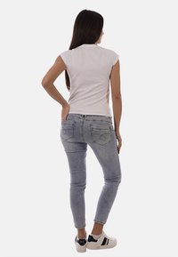 Donna con capelli lunghi e scuri vista di spalle, indossa una canottiera bianca, jeans azzurri e sneakers bianche con strisce nere.