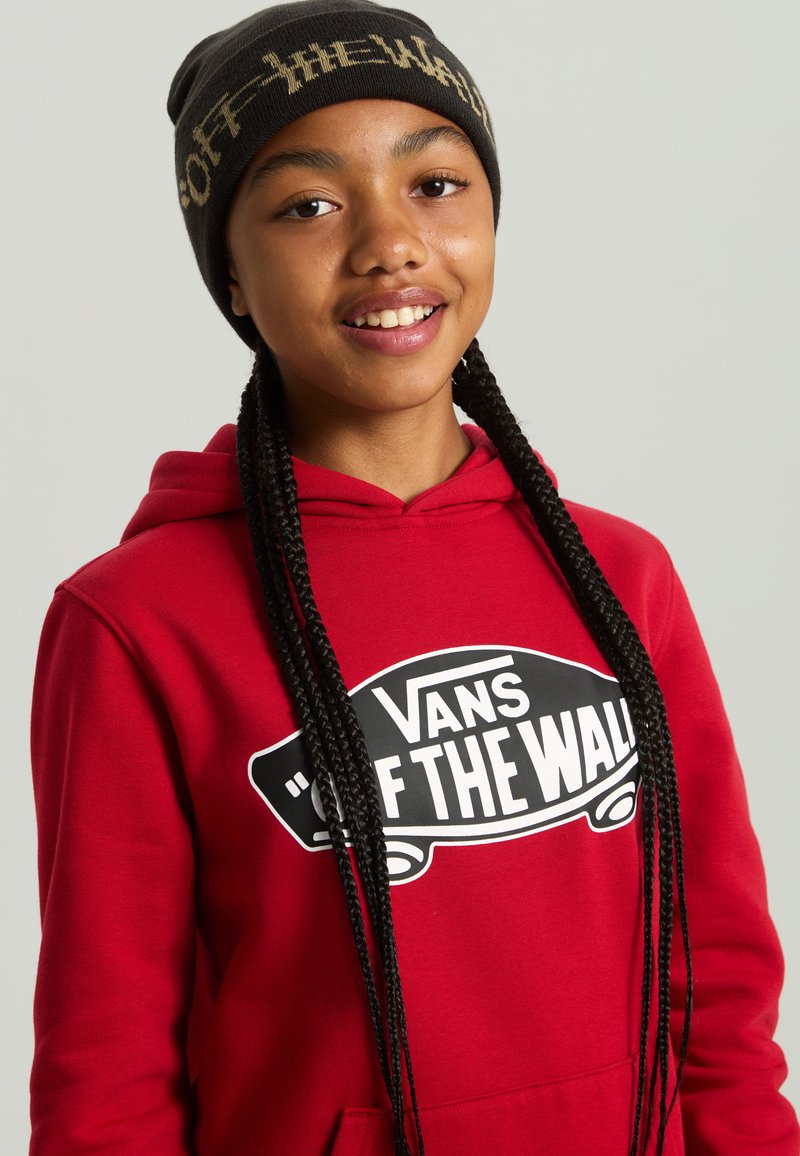 Rød hættetrøje med "VANS OFF THE WALL" grafik i sort, parret med en sort beanie med hvid tekst. Lange flettede hår er synlige.