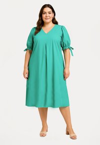 Türkisfarvet kjole lavet af blødt stof, med V-hals, korte pufærmer med bånd og en plisseret front. Knælangt design.