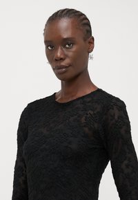 Svart langermet topp med blomstermotiver i blonde. Stoffet er gjennomsiktig med en teksturert overflate, og har rund halsutskjæring.