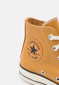 Tenisky typu high-top Converse All Star v texturovaném hořčicově žlutém materiálu. Obsahují bílé šití, kovové očky a černý nášivku s hvězdou.