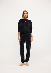 Crni velours komplet za opuštanje koji se sastoji od majice dugih rukava i trenirki. Zapaženi crveni logo na oba komada. Mekana tekstura, opušten kroj.