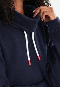 Felpa blu navy con colletto alto a scaldacollo, caratterizzata da lacci bianchi con punte rosse e una trama a coste. Design casual e confortevole.