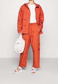 Orange vindjacka med fickor, matchande cargo-byxor och vita sandaler. En krämfärgad ryggsäck hålls i ena handen. Minimalistisk design.