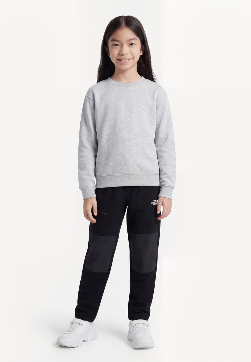 Lány áll, világosszürke pulóvert, térdén foltos fekete nadrágot és fehér tornacipőt visel, egyszerű fehér háttér előtt.