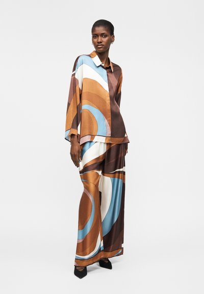 Γυναίκα με φαρδιά πουκάμισο και παντελόνι με φαρδιά μπατζάκια, με αφηρημένα στροβιλισμένα σχέδια σε καφέ, μπλε, λευκό και ταμπέ αποχρώσεις, στέκεται με ουδέτερη έκφραση.