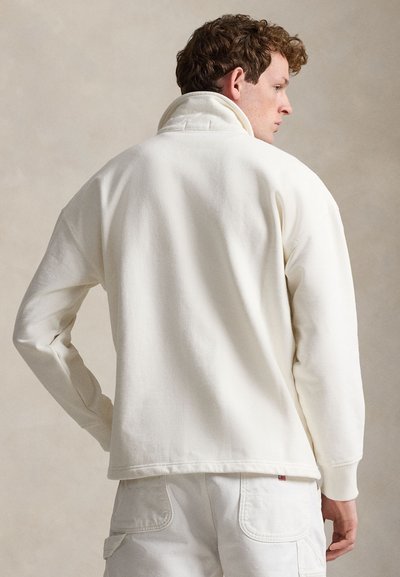 Άνδρας με σγουρά μαλλιά που φοράει ένα εκρού φλις πουλόβερ με ψηλό γιακά και λευκό παντελόνι cargo, γυρισμένος προς τα πίσω μπροστά σε ουδέτερο φόντο.