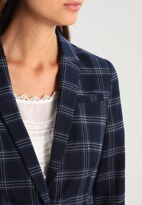 Femme portant un blazer à carreaux bleu marine sur une blouse blanche bordée de dentelle, tournée vers la droite, sur un fond gris clair.