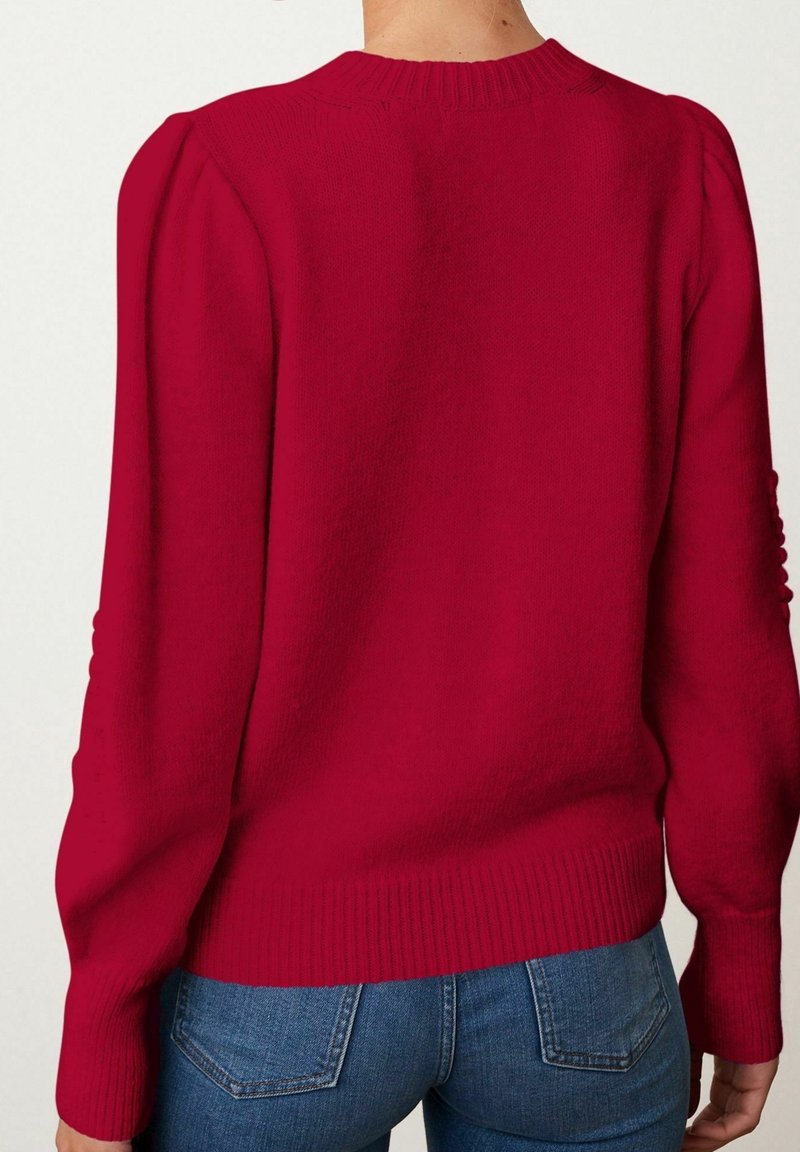 Femme portant un pull tricoté rouge vif à manches longues avec des épaules bouffantes, associé à un jean en denim bleu, vue de dos.