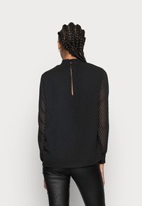 Blouse noire à manches longues avec un motif chevrons texturé, col montant et détail dos en forme d'œillet. Portée avec un pantalon noir.