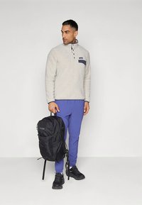 Pulôver de lã polar cinza claro com um bolso no peito azul-marinho, combinado com calças azuis e uma mochila preta com logótipo da marca e vários bolsos.