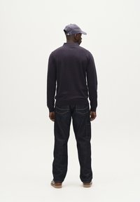 Tengerészkék kötött pulóver gallérral, sötét farmernadrággal párosítva, amely oldalzsebet és látható varrást tartalmaz. A megjelenést egy világos sapka teszi teljessé.